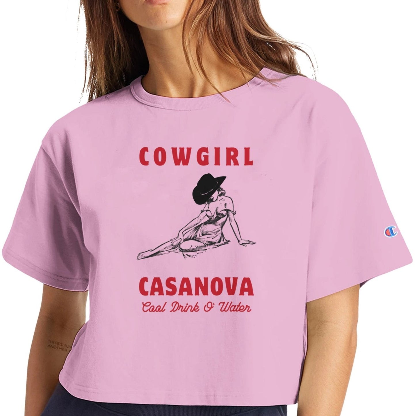 Cropped shirt in pink with cowgirl graphic leaning on her hand, her face covered by a hat. Shirt text reads "Cowgirl Casanova" In bold red letters above and below graphic. Below in thin red cursive it says "Cool drink o' water"