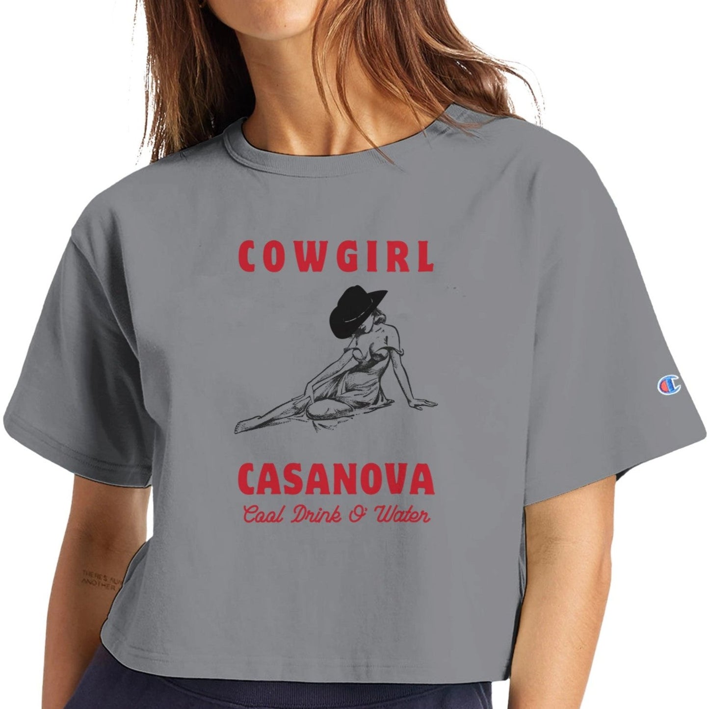 Cropped shirt in gray with cowgirl graphic leaning on her hand, her face covered by a hat. Shirt text reads "Cowgirl Casanova" In bold red letters above and below graphic. Below in thin red cursive it says "Cool drink o' water"