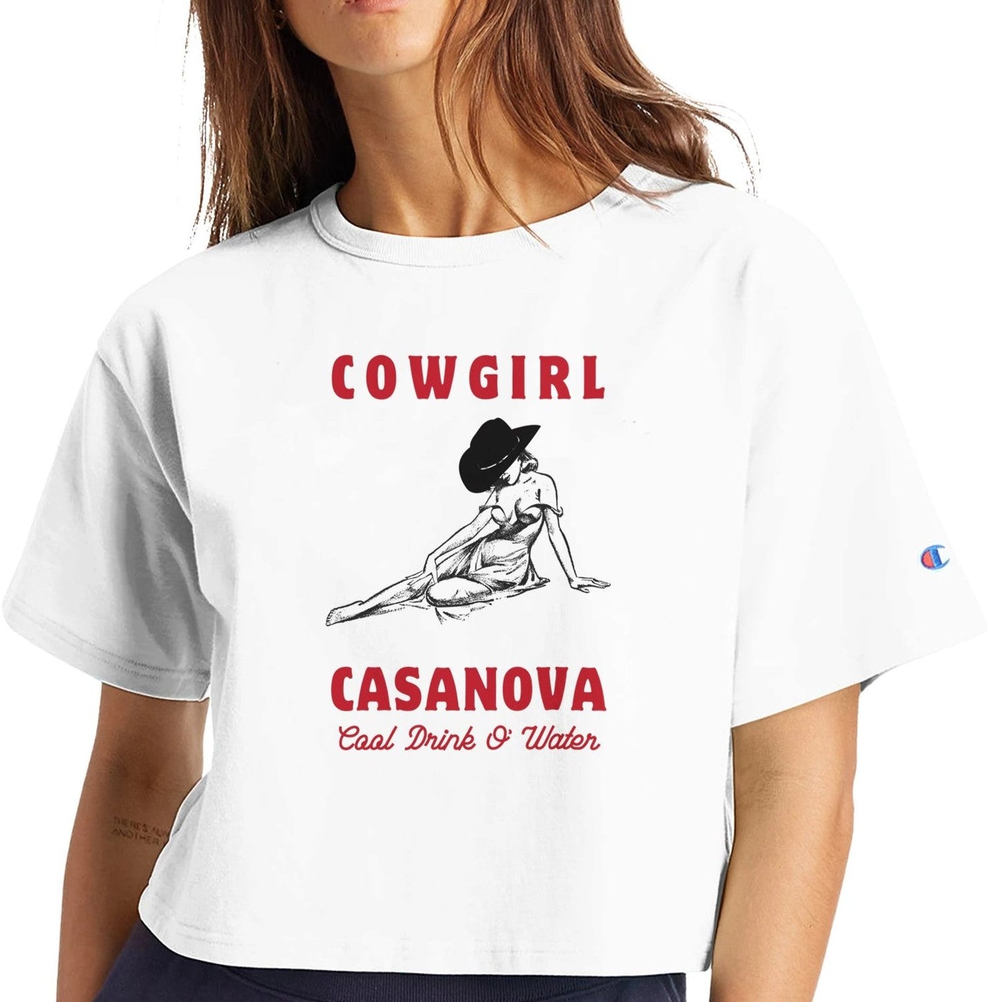 Cropped shirt in white with cowgirl graphic leaning on her hand, her face covered by a hat. Shirt text reads "Cowgirl Casanova" In bold red letters above and below graphic. Below in thin red cursive it says "Cool drink o' water"